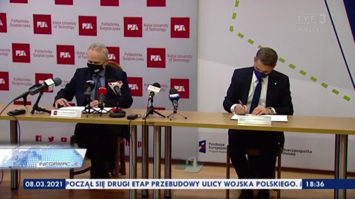 Zrzut ekranu z materiału telewizyjnego TVP3 Kielce. Przy stole siedzi dwóch mężczyzn i podpisuje dokumenty. Na stołach stoją mikrofony dziennikarskie. Z tyłu widać roll-upy partycypantów umowy.