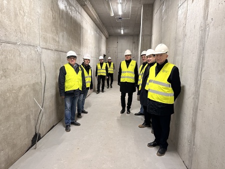  Ośmiu mężczyzn w żółtych kamizelkach roboczych i w białych kaskach na głowach stoi w wybetonowanym korytarzu znajdującym się w stanie budowlanym surowym.